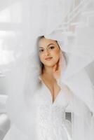 porträtt av en anbud brud i en bröllop klänning och en skön frisyr under en slöja i henne rum från de morgon- foto
