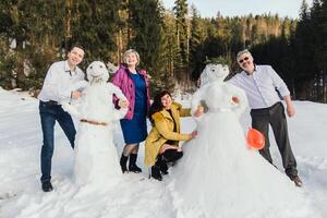 Lycklig senior vänner har roligt framställning en snögubbe tillsammans mot de bakgrund av en tall skog. vänner foto