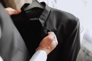 de man innehar hans svart jacka på en galge i hans händer. de brudgum är framställning för de bröllop ceremoni. detaljerad närbild Foto av händer
