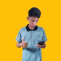 upphetsad asiatisk man bär grå t-shirt pekande till kopia Plats Nästa till innehav smart telefon, isolerat på gul bakgrund. foto