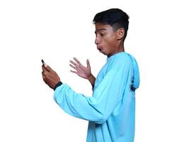 ung asiatisk man överraskad ser på smart telefon bär blå t-shirt, isolerat vit bakgrund. foto