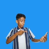 ung asiatisk man överraskad ser på smart telefon, bär färgrik t-shirt, isolerat blå bakgrund. foto