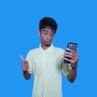ung asiatisk man överraskad ser på smart telefon bär gul t-shirt, blå bakgrund. foto