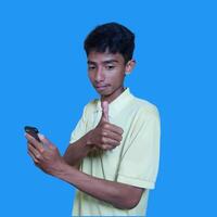 ung asiatisk man överraskad ser på smart telefon, bär gul t-shirt, isolerat blå bakgrund. foto