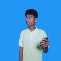 ung asiatisk man överraskad ser på smart telefon isolerat blå bakgrund. bär en gul t-shirt foto
