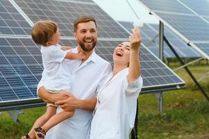 sol- energi begrepp. en ung, Lycklig familj är stående nära sol- paneler och innehav ett elektrisk ljus Glödlampa. foto