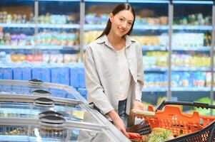 ung leende Lycklig kvinna 20s i tillfällig kläder handla på supermarket Lagra med matvaror vagn foto