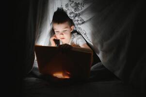 läsning bok och använder sig av ficklampa. ung pojke i tillfällig kläder liggande ner nära tält på kväll tid foto