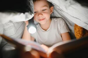 pojke med ficklampa läsning bok under filt på Hem. foto
