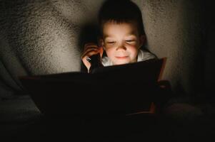 porträtt av söt liten pojke läsning i säng med ficklampa i mörk rum, njuter sagor. foto