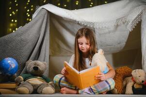 en Söt liten flicka läsning en bok på de golv under de lampa. barn och utbildning foto