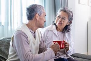 par av friska asiatisk senior far och mor Sammanträde på soffa med Lycklig leende på pensionering Hem dricka varm te till fira deras Semester tillsammans för äldre vård och utgifterna värdefullt tid foto