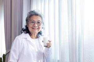 senior asiatisk lady dricka glas av mjölk medan stående förbi de fönster med kopia Plats för kalcium öka och friska mejeri produkt konsumtion begrepp foto