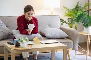 en ung asiatisk kvinna sitter på en soffa i henne Hem, känsla orolig och frustrerad handla om henne en gång i månaden utgifter. olika verktyg räkningar. foto