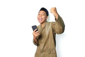 glad asiatisk muslim man bär islamic klänning visa vinnande gest med innehav mobil telefon isolerat på whtie bakgrund foto