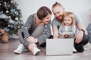 vacker ung mamma, pappa och deras dotter använder laptop, pratar och ler när de sitter i soffan hemma foto
