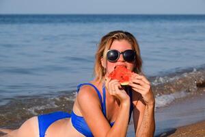 de blond i solglasögon lögner på de strand nära de vågor och äter en vattenmelon. en saftig vattenmelon i de händer av en kvinna. foto