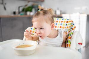 söt bebis flicka litet barn Sammanträde i de hög stol och äter henne lunch soppa på Hem kök. foto