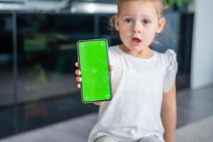 liten flicka använder sig av en telefon med en grön skärm. en liten flicka är Sammanträde och innehav en smartphone med en Chromakey foto