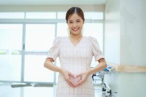 Lycklig och upphetsad gravid kvinna framställning hjärta form på henne mage på sjukhus foto