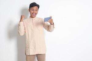 porträtt av upphetsad asiatisk muslim man i koko skjorta innehav mobil telefon och spelar spel på hans smartphone, höjning händer, fira Framgång, bli en vinnare. isolerat bild på vit bakgrund foto