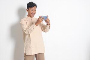 porträtt av ung upphetsad asiatisk muslim man i koko skjorta innehav mobil telefon och spelar spel på hans smartphone. isolerat bild på vit bakgrund foto