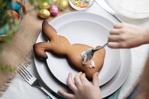 närbild av kanin formad pepparkaka kaka. händer av barn dekorera kex med glasyr på tallrik. påsk Semester traditioner foto