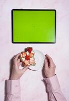 kvinna innehav mjuk ost och jordgubb smörgås nära grön skärm läsplatta foto