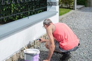 en medelålders man målarfärger en staket med vit måla med en borsta, reparationer de skadad yta foto