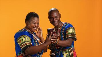 traditionell afrikansk amerikan par bläddring webbplatser på smartphone, kontroll foton på social media app och skrattande på memes. glad människor i färgrik klädsel ser på bilder uppkopplad.