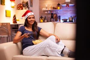 vuxen person bär röd santa hatt liggande på soffa tittar på jul komedi film under jultid i xmas dekorerad kök. kvinna njuter vinter- säsong fira x-mas Semester foto