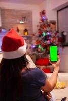 kvinna innehav grön skärm falsk upp krom nyckel telefon med isolerat visa Sammanträde bekväm på soffa i xmas dekorerad kök. person fira jul Semester njuter vinter- säsong foto