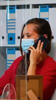 kvinna talande på telefon bär medicinsk ansikte mask i de kontor respekterar social avståndstagande. frilansare arbetssätt i ny vanligt arbetsplats chattar med avlägset team tala på smartphone foto