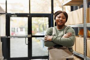 leende lager chef stående med ärm korsade, Framställ med förtroende i lager. afrikansk amerikan anställd bär industriell övergripande medan arbetssätt på kunder uppkopplad order foto