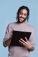 leende arab man innehav Urklipp och skrivning planen checklista med glad ansiktsbehandling uttryck. sorglös ung person bär tillfällig kläder tar anteckningar och fyllning Ansökan foto