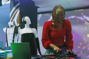 leende afrikansk amerikan kvinna blandning ljud med dj station på skede medan utför i nattklubb. Lycklig musiker använder sig av elektronisk musik mixer trösta på diskotek fest i klubb foto