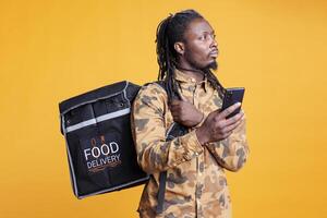 afrikansk amerikan kurir ser på kund adress på smartphone ta ut app innan leverera beställa under lunch tid. deliveryman bärande termisk ryggsäck full med hämtmat mat över foto