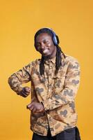 afrikansk amerikan man lyssnande musik använder sig av hörlurar, dans ensam i studio över gul bakgrund. porträtt av leende ung vuxen njuter glad Spellista, har roligt på låt rytm foto