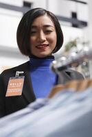 mode boutique leende asiatisk kvinna säljare med bricka granskning formell shirts på visa kuggstång. avdelning handla Centrum Kläder Lagra assistent hantera plagg lager foto