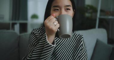 porträtt av avkopplande ung kvinna innehav kopp njut av lukt kaffe eller te och dricka med lycka i morgon- medan sitta på soffa i levande rum, gratis tid, ta ha sönder i hem, ler foto