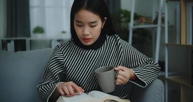 porträtt av Lycklig ung asiatisk kvinna dricka morgon- kaffe eller te och läsning i levande rum på Hem på helgen. fritid och livsstil, gratis tid foto