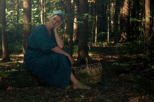 ung kvinna i folk dräng kläder med en korg för samlar vild växter sitter på en logga i de barr- skog foto