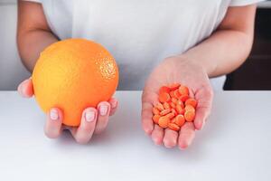 de flicka är innehav en mogen orange i ett hand, och vitamin c tabletter i de Övrig hand. val mellan naturlig och syntetisk sätt av hälsa vård. friska livsstil begrepp. närbild. foto