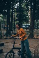 Lycklig ung asiatisk kvinna medan ridning en cykel i en stad parkera. hon log använder sig av de cykel av transport. miljömässigt vänlig begrepp. foto