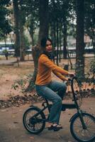 Lycklig ung asiatisk kvinna medan ridning en cykel i en stad parkera. hon log använder sig av de cykel av transport. miljömässigt vänlig begrepp. foto