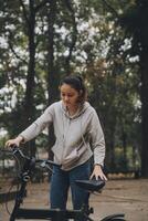 Lycklig asiatisk ung kvinna promenad och rida cykel i parkera, gata stad henne leende använder sig av cykel av transport, eco vänlig, människor livsstil begrepp. foto