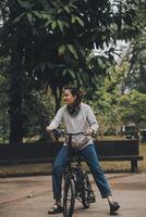 Lycklig asiatisk ung kvinna promenad och rida cykel i parkera, gata stad henne leende använder sig av cykel av transport, eco vänlig, människor livsstil begrepp. foto