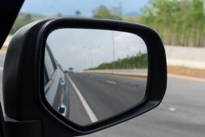 spegel se av bil resa på de asfalt väg. bakgrund på främre med hastighet av asfalt väg och istället av en vid vägkanten barriär och sudd träd. foto