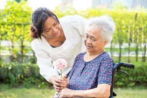 vårdgivare prata och hjälp asiatisk äldre kvinna innehav blomma, leende och Lycklig i de solig trädgård. foto