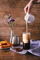 kvinnors hand med gräddkanna häller mjölk i glas med kaffe. kanel pinnar, hemlagad småkakor och knippa av vild på en trä- bakgrund. frukost begrepp. foto